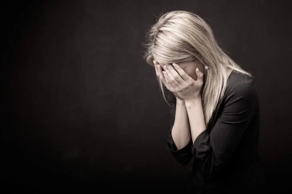 sur fond noir et ambiance sans couleur, une femme blonde a les mains qui lui cachent le visage. elle a le dos légèrement recourbée et est habillée de noir.