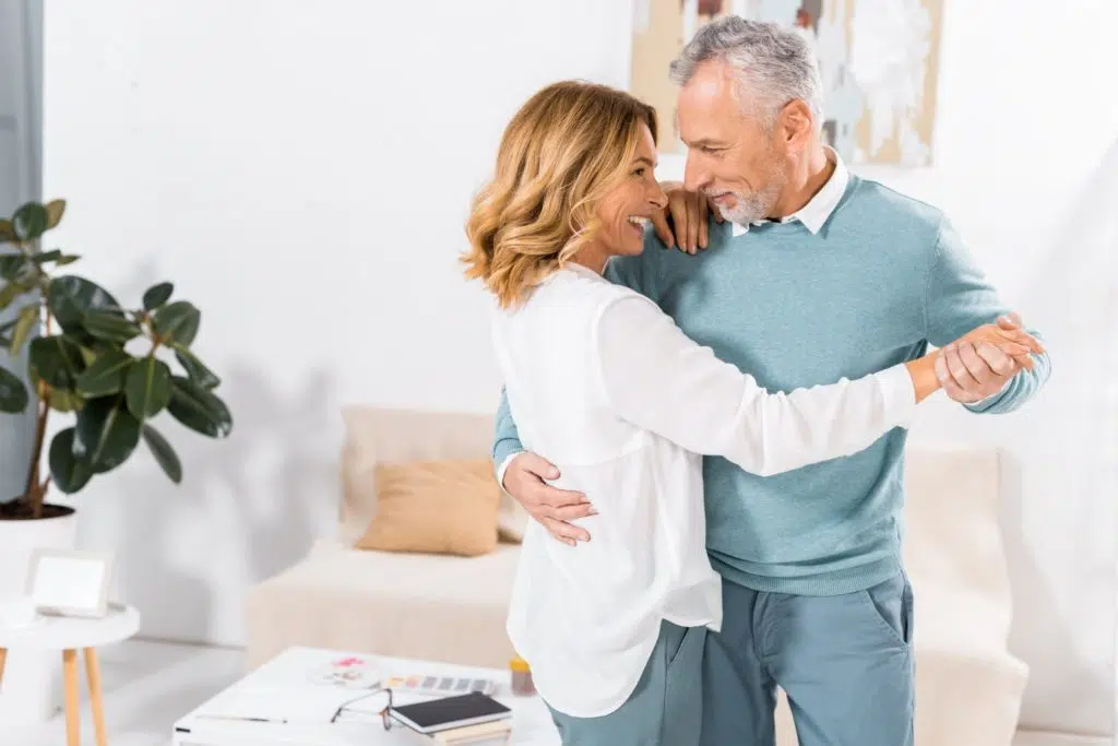 un couple de personnes mûres danse dans un salon d'intérieur en se souriant l'un l'autre.