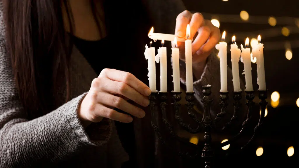 des mains de jeune femme qui tient une bougie pour allumer d'autres bijoux sur un chandelier sur un fond sombre et des ponts de lumières en arrière plan. 