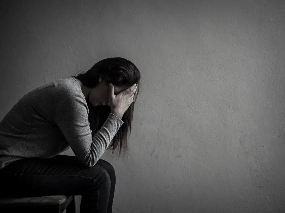 photo en noir et blanc d'une femme assise qui se tient la tête baissée devant un mur gris. 